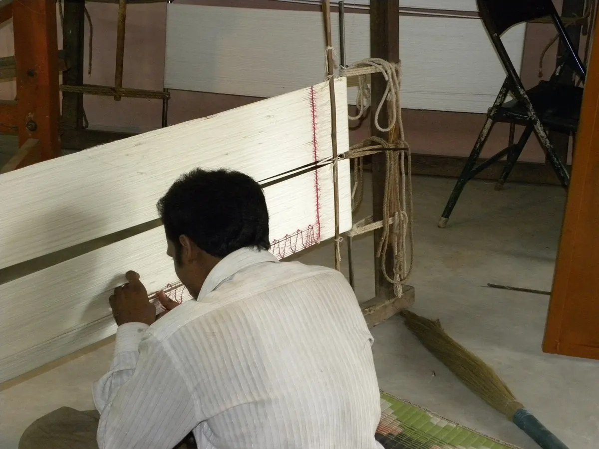 A man kneels at a long frame of stretched white yarn, working along it by hand.