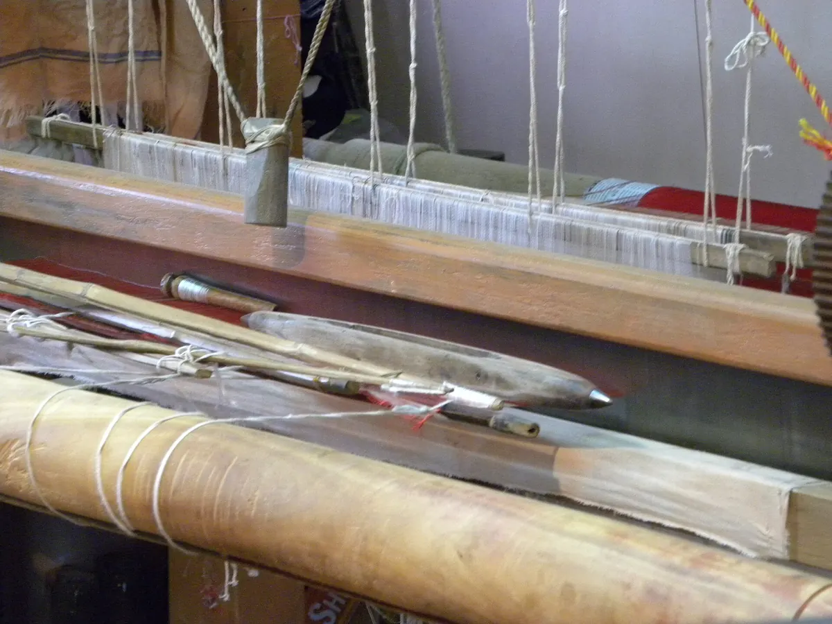 Wooden shuttles resting on the beam of a handloom, white warp threads above.
