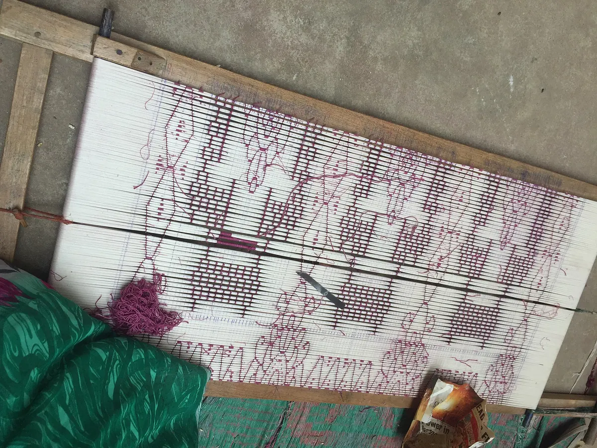 White yarn stretched on a wooden frame, a bandha design of fish, squares and spires marked out on it in purple thread.