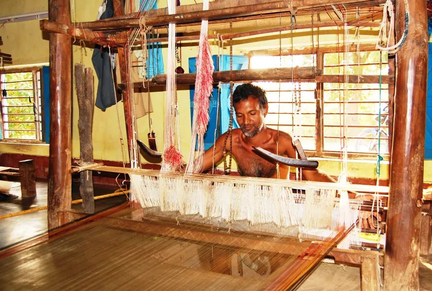 A weaver at a wooden handloom, coloured yarn hanging from the frame above.