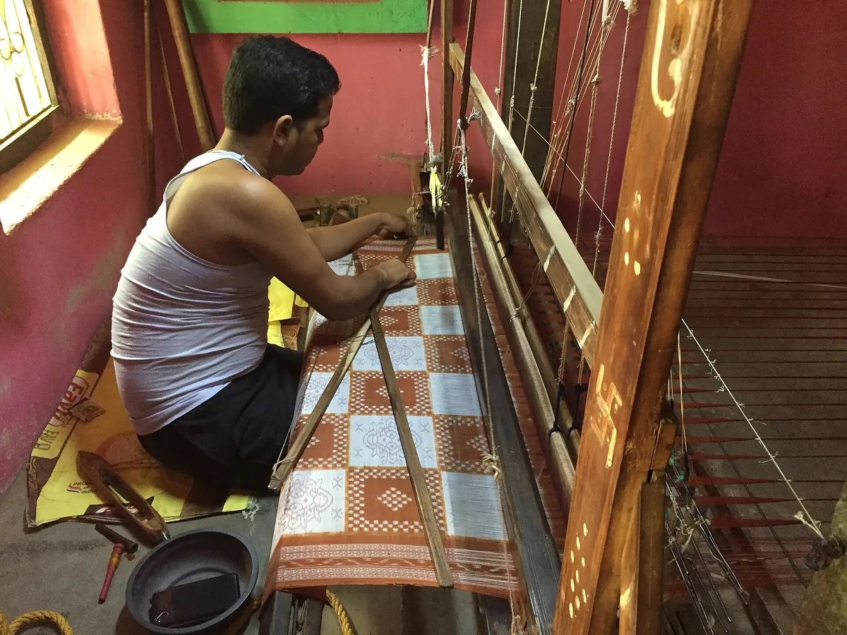 A weaver sits at a loom in a red-walled room, weaving a saree of brown and white squares with small motifs.